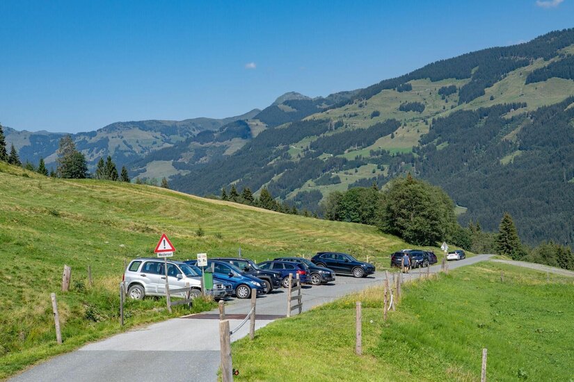 Parkende Autos am Rand einer asphaltierten Bergstraße vor bewaldeten Hängen.