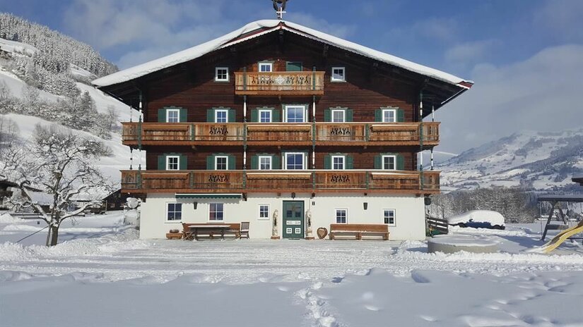Winteransicht Angererhof Großes, mehrstöckiges Holzhaus mit Balkonen in einer verschneiten Winterlandschaft unter blauem Himmel.