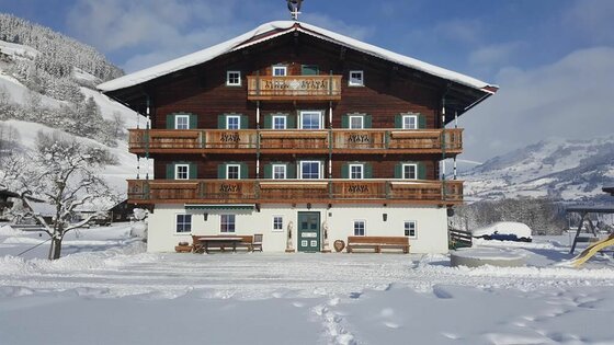 Winteransicht Angererhof Großes, mehrstöckiges Holzhaus mit Balkonen in einer verschneiten Winterlandschaft unter blauem Himmel.