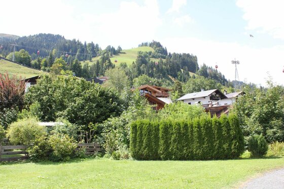 Aussicht auf Kitzbüheler Berghang Blick über eine Wiese und Hecke auf Häuser vor einem bewaldeten Berghang mit Gondelbahn.