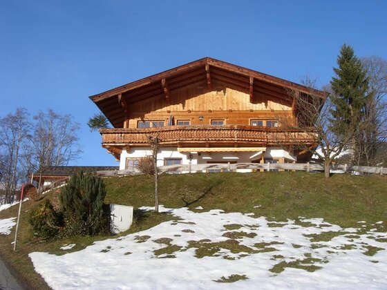 Zweistöckiges Holzhaus mit Balkon auf einem Grashügel mit Schneeresten unter blauem Himmel.