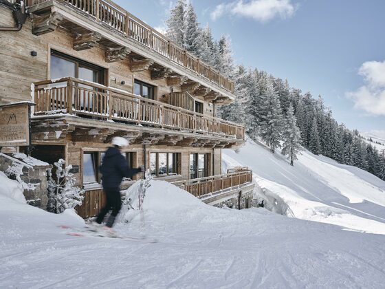 Mehrstöckige Holzlodge an einer verschneiten Skipiste mit einem Skifahrer im Vordergrund.
