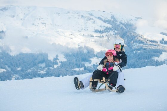 Zwei Personen fahren gemeinsam auf einem Holzschlitten eine verschneite Piste hinunter.
