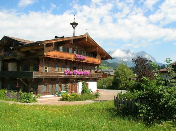 Neuhaus Ein traditionelles Holzgebäude mit Blumenbalkonen in einer ländlichen Umgebung. Im Hintergrund sind grüne Felder und leichte Berge zu sehen.