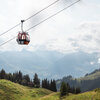 Die rote Hahnenkammbahn schwebt über die grünen Hänge in Kitzbühel, umgeben von Bergen und einem bewölkten Himmel.