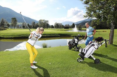 Eine Frau beim Golfabschlag auf einem grünen Golfplatz mit Wasserhindernis und Bergen im Hintergrund.
