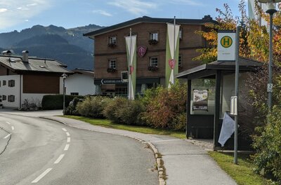 Bushaltestelle Tischlerwirt Bushaltestelle an einer Straße vor einem mehrstöckigen Holzgebäude mit Bergen im Hintergrund.