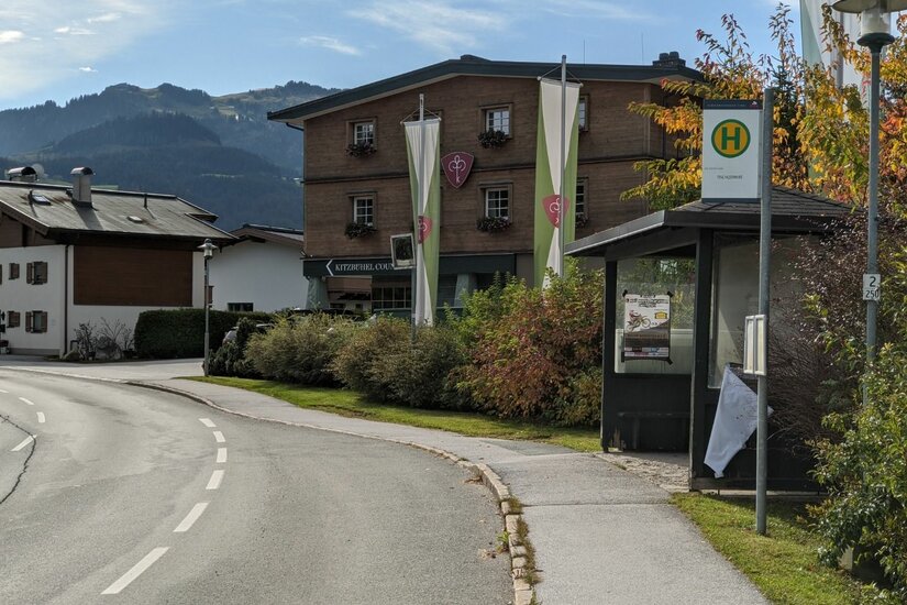 Bushaltestelle an einer Straße vor einem mehrstöckigen Holzgebäude mit Bergen im Hintergrund.