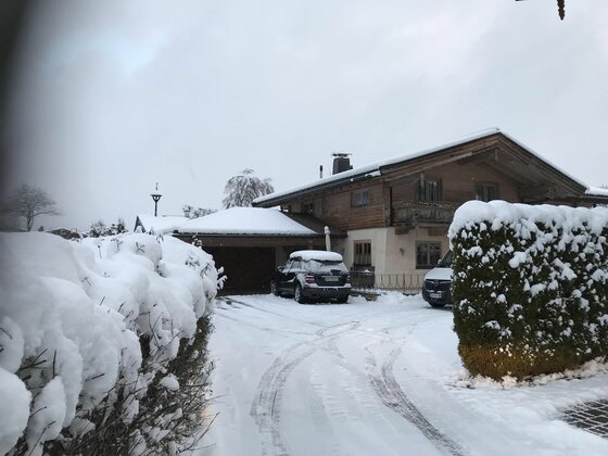Egger Studio Hornblick im Winter Ein schneebedecktes Holzhaus mit Garage, davor parkende Autos und eine verschneite Hecke unter grauem Himmel.