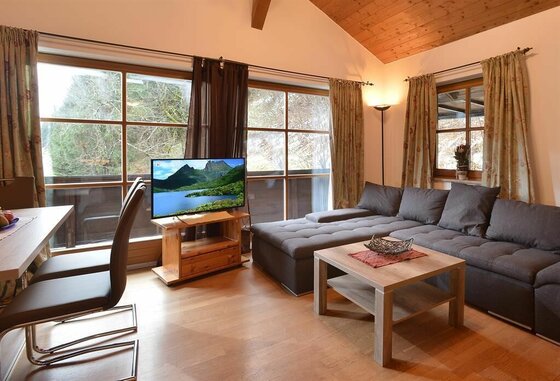 Wohnzimmer im Appartement Holzleitner Wohnzimmer mit großem Ecksofa, Couchtisch, Fernseher und Fensterfront mit Blick ins Grüne.