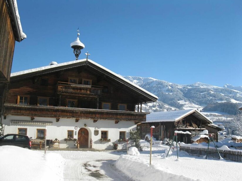 Traditionelles Bauernhaus aus Holz und weißem Putz in einer verschneiten Gebirgslandschaft unter blauem Himmel.