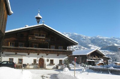 Traditionelles Bauernhaus aus Holz und weißem Putz in einer verschneiten Gebirgslandschaft unter blauem Himmel.