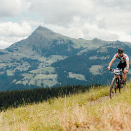 In Kitzbühel fährt ein Mountainbiker auf schmalem Pfad durch grüne Wiese mit Bergkulisse und Wolken.