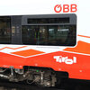 Ein ÖBB-Zug mit einem modernen Design und einem Mitarbeiter in Warnkleidung, der an einem belebten Bahnhof steht.