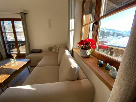 Wohnbereich im Haus Ingrid Wohnzimmer mit beigem Sofa, Holztisch und Fenster mit Blick auf verschneite Berge.