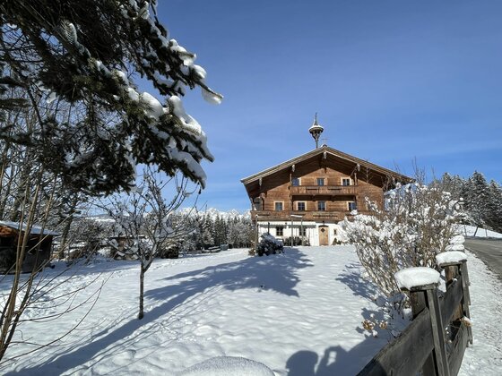 Ein traditionelles Holzhaus mit Glockenturm in einer verschneiten Winterlandschaft unter blauem Himmel.