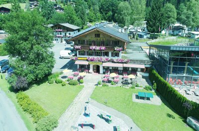 Ein malerisches Hotel umgeben von grünen Bäumen und bunten Blumen. Terassen mit Tischen und Stühlen laden zum Entspannen ein.