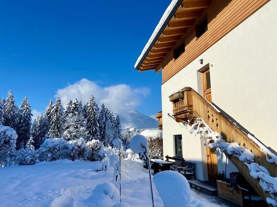 Ein hübsches Holzhaus im Schnee mit einem Balkon. Im Hintergrund sind schneebedeckte Bäume und ein blauer Himmel zu sehen.