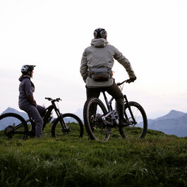 Zwei Mountainbiker stehen bei Sonnenaufgang auf einem Hügel und blicken über die umliegenden Berge am Hahnenkamm in Kitzbühel