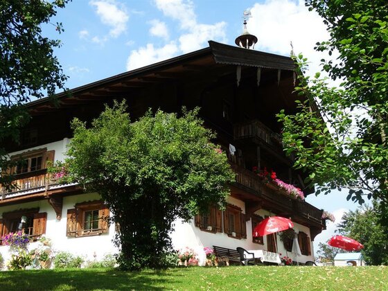 Ansicht Plattenhof Alpenländisches Bauernhaus mit Holzbalkonen, Blumenkasten und Sonnenschirm auf einer Wiese.