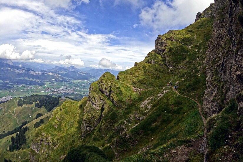 Schmaler Wanderpfad an einem steilen, grasbewachsenen Berghang mit Blick ins Tal.