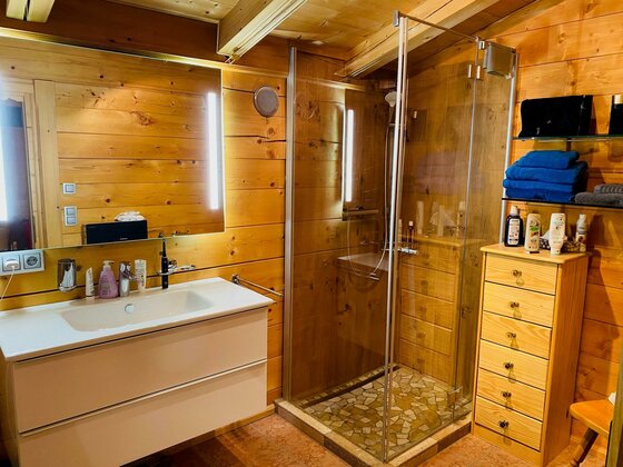 Badezimmer mit Holzwaenden, weißem Waschtisch, Glasdusche und einer Kommode aus Naturholz.