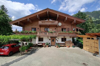 Haus Berger Alpines Holzhaus mit Blumenbalkon, gepflastertem Vorplatz und einem roten Auto unter einer Pergola.