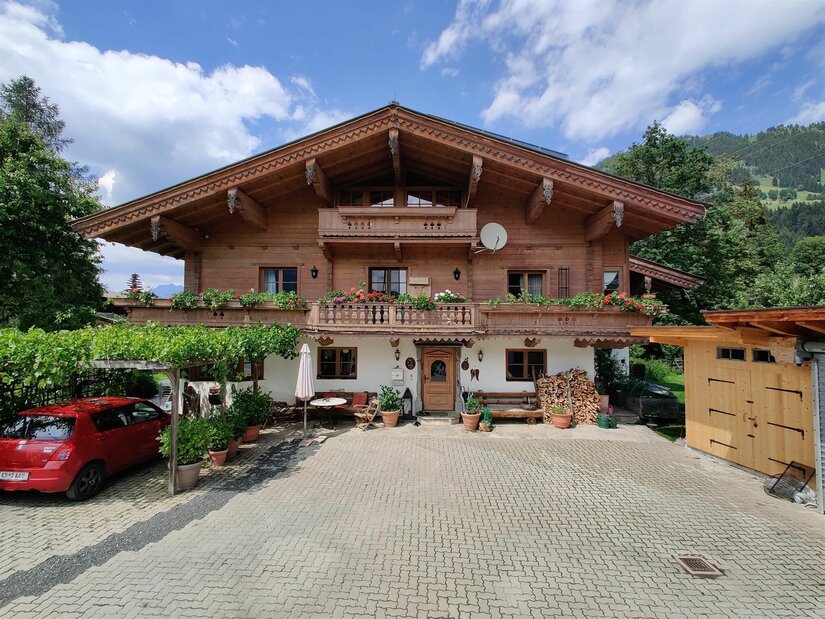 Alpines Holzhaus mit Blumenbalkon, gepflastertem Vorplatz und einem roten Auto unter einer Pergola.