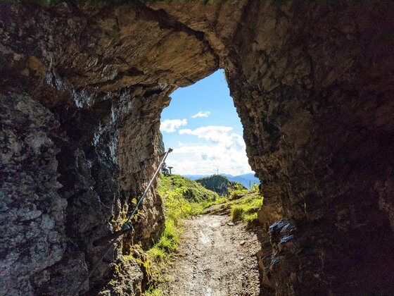 Blick aus einem dunklen Felstunnel auf einen sonnigen Bergpfad mit Drahtseilsicherung unter blauem Himmel.