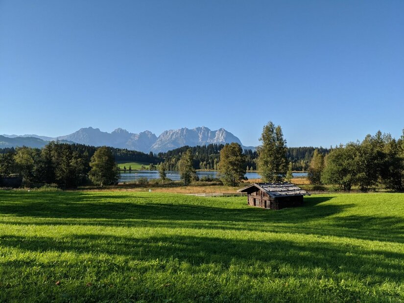 Schwarzsee mit Kaisergebirge Grüne Wiese mit kleiner Holzhütte vor einem See und Gebirgskette unter blauem Himmel.