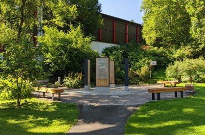 Gepflasterter Platz mit hölzernen Infostelen und Sitzbänken inmitten von Bäumen und Sträuchern.