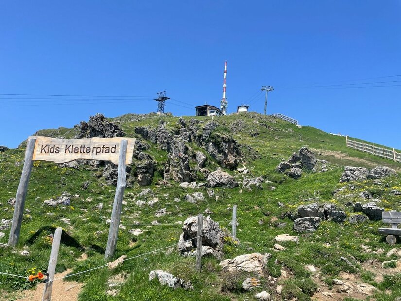 Holzschild mit der Aufschrift Kids Kletterpfad auf einem grasbewachsenen Berghang unter blauem Himmel.