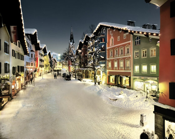 Verschneite Fußgängerzone in Kitzbühel bei Nacht mit beleuchteten bunten Häuserfassaden und Kirchturm.
