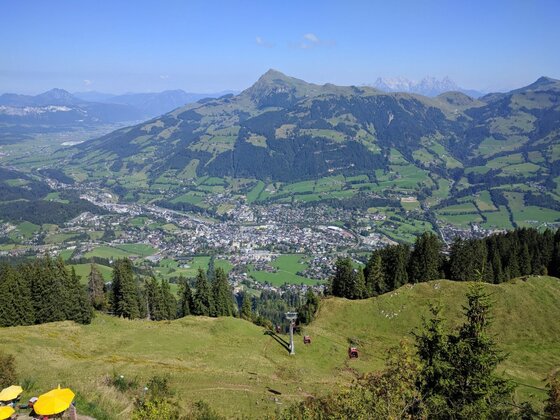 Blick von einem Grashang auf eine Stadt im Tal und gegenüberliegende bewaldete Berge unter blauem Himmel.