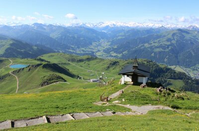Eine malerische Berglandschaft mit grünen Wiesen und schneebedeckten Gipfeln im Hintergrund. Im Vordergrund steht eine kleine Kapelle auf einem Hügel.