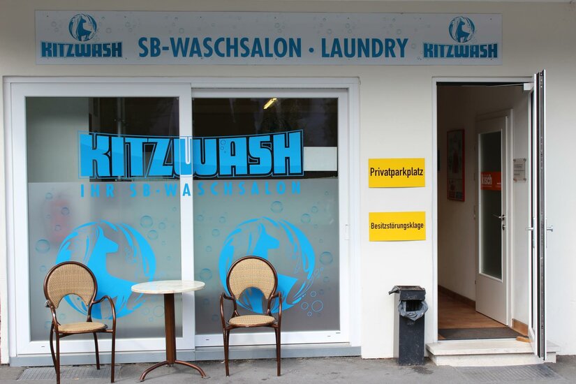 Kitz Wash Ein Waschsalon mit der Aufschrift "Kitzwash". Vor dem Eingang stehen zwei Stühle und ein kleiner Tisch.