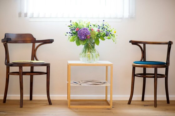 Stadtpraxis Zwei Holzstühle stehen neben einem kleinen Tisch. Auf dem Tisch steht eine Vase mit bunten Blumen.