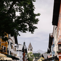 Eine malerische Straße mit bunten Häusern und einem hohen Kirchturm, umgeben von üppigem Grün und sanften Bergen im Hintergrund.