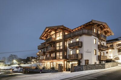 Mehrstöckiges alpines Gebäude mit Holzfassade und beleuchteten Fenstern in der Dämmerung bei Schnee.
