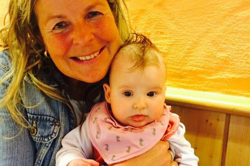 Christa Blaschke Eine lächelnde Frau hält ein Baby in den Armen. Der Hintergrund ist orangefarben, was eine freundliche Atmosphäre schafft.