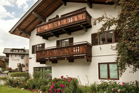 Alpines Wohnhaus mit weißen Wänden, zwei Holzbalkonen und Blumenbeeten im Vordergrund.