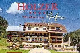 Hotel Holzer im Sommer Mehrstöckiges Hotelgebäude mit Balkonen und Café-Restaurant vor bewaldetem Hang unter blauem Himmel.