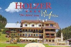 Hotel Holzer im Sommer Mehrstöckiges Hotelgebäude mit Balkonen und Café-Restaurant vor bewaldetem Hang unter blauem Himmel.
