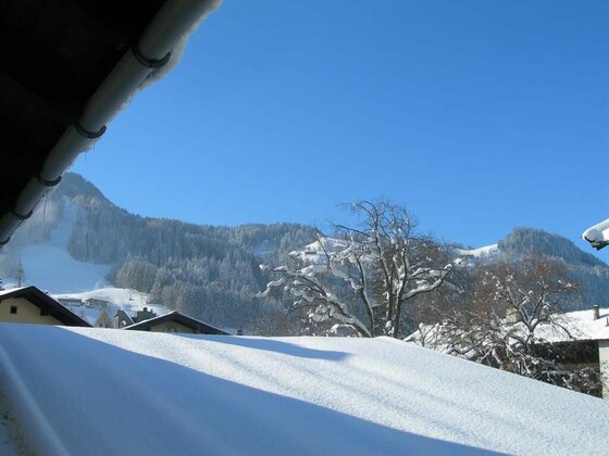 Aussicht Winter Apartment Gaby Blick über schneebedeckte Dächer auf bewaldete Berge unter blauem Himmel.