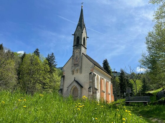 Kapelle bei der Kelchalm Kleine Kapelle mit spitzem Turm auf einer grünen Wiese mit gelben Blumen vor Waldhintergrund.