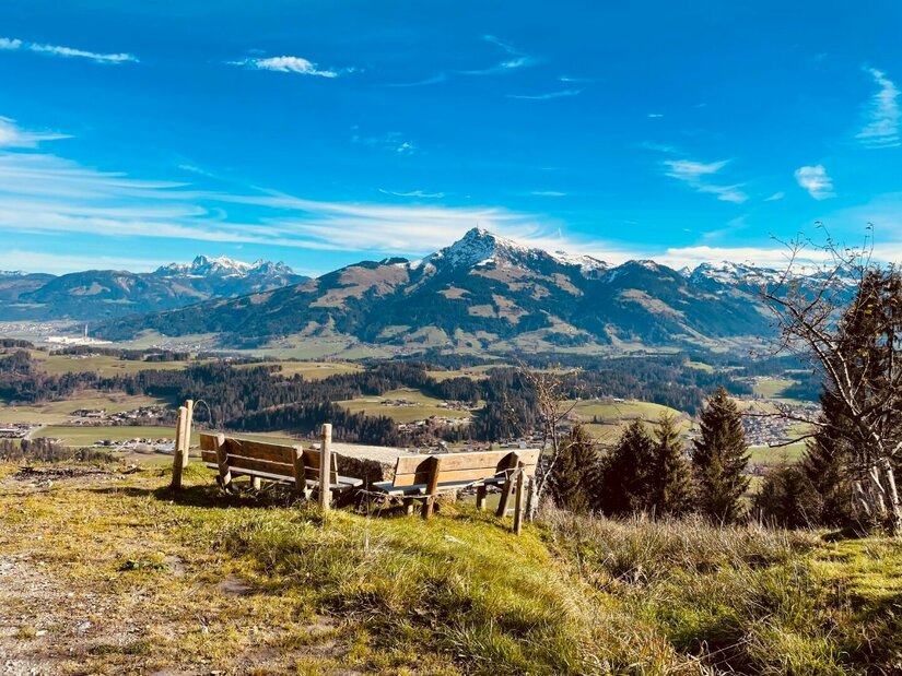 Rastplatz am Höhenweg Reith Holzsitzgruppe auf einer Wiese mit Blick auf ein Gebirgspanorama unter blauem Himmel.