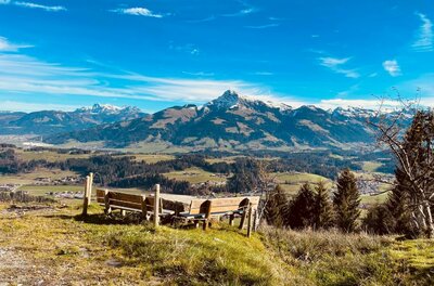Rastplatz am Höhenweg Reith Holzsitzgruppe auf einer Wiese mit Blick auf ein Gebirgspanorama unter blauem Himmel.