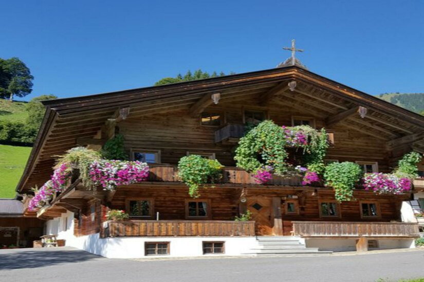Hof Köglern Ein traditionelles Chalet mit detaillierter Holzarchitektur. Das Haus ist mit bunten Blumen und Pflanzen geschmückt, umgeben von grünen Bergen und einem blauen Himmel.