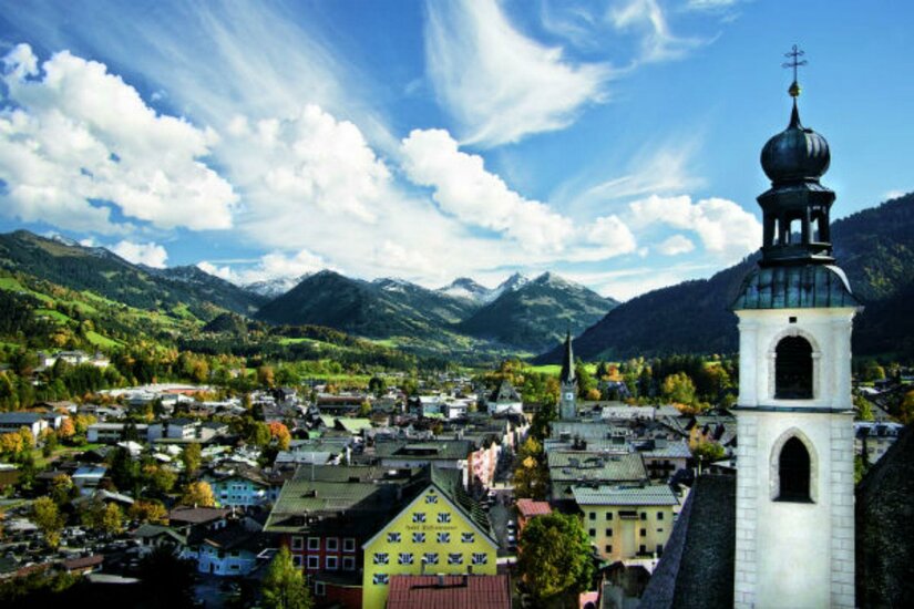 Eine malerische Landschaft mit einem Blick auf ein kleines Dorf umgeben von Bergen. Die Wolken am blauen Himmel verleihen der Szene eine friedliche Atmosphäre.