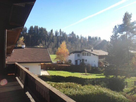 Blick von einem Holzbalkon auf Wohnhäuser, eine Wiese und einen bewaldeten Hügel unter blauem Himmel.